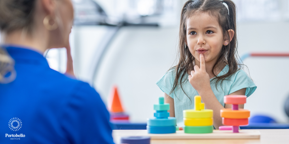a young girl sitting across from an adult with her finger to her mouth making a sound