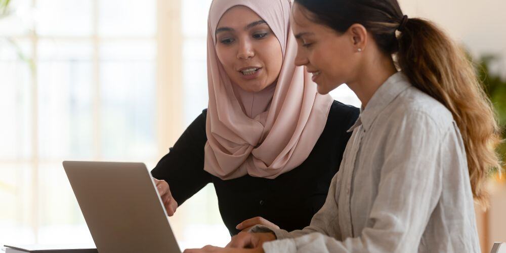 One woman appears supporting another woman sitting at a table looking at a laptop