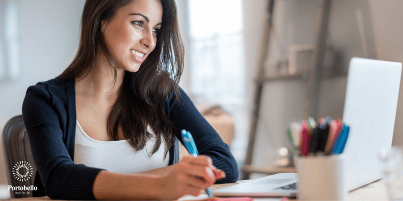 smiling woman looking at laptop screen holding a pen and taking notes 