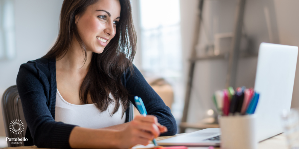 smiling woman looking at laptop screen holding a pen and taking notes 