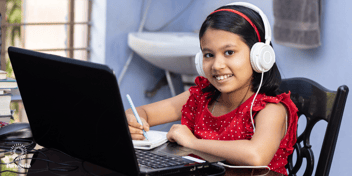 A young girl sitting at a laptop with headphones on smiling