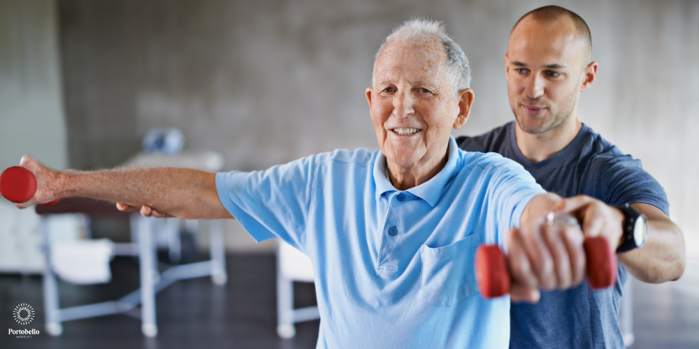 a physiotherapist working with an older man supporting his arms while he lifts light weights smiling