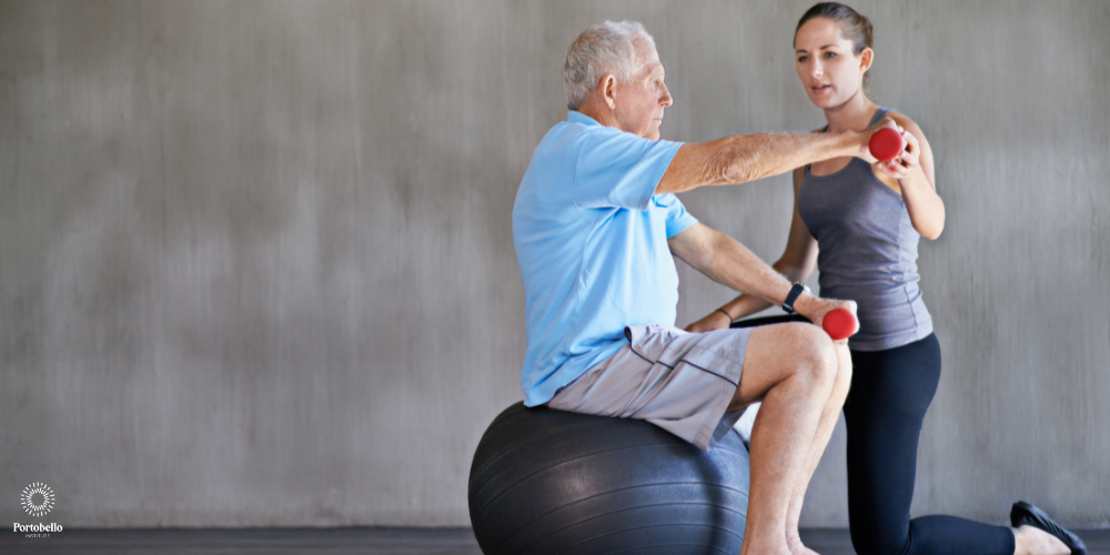 sports therapist assisting an older man to hold a small weight while sitting on a ball