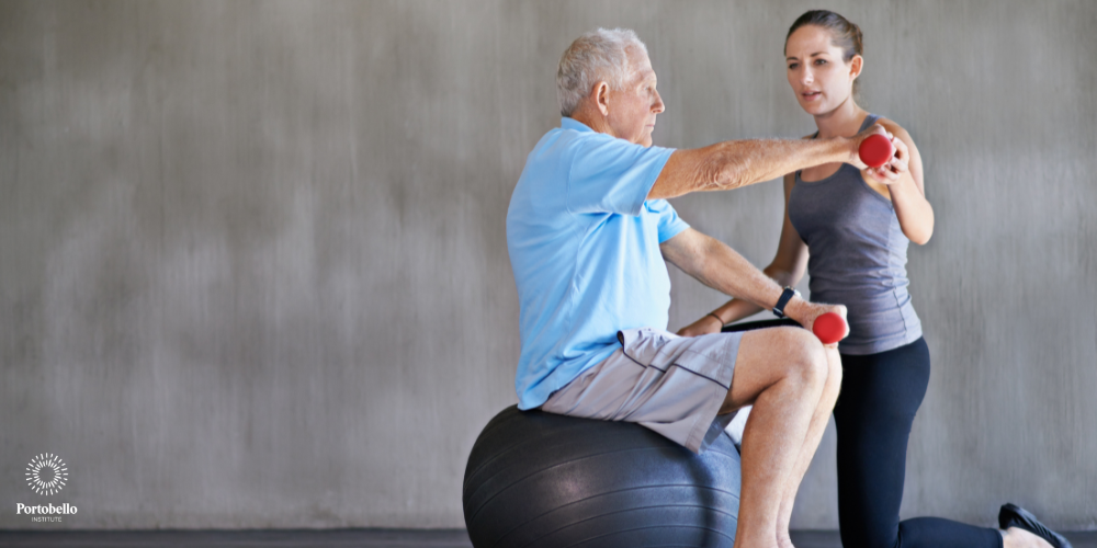 physiotherapist supports a older man on an exercise ball to lift weights