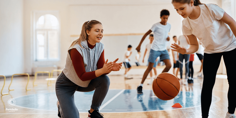 A PE teacher clapping as a young student bounces a basketball